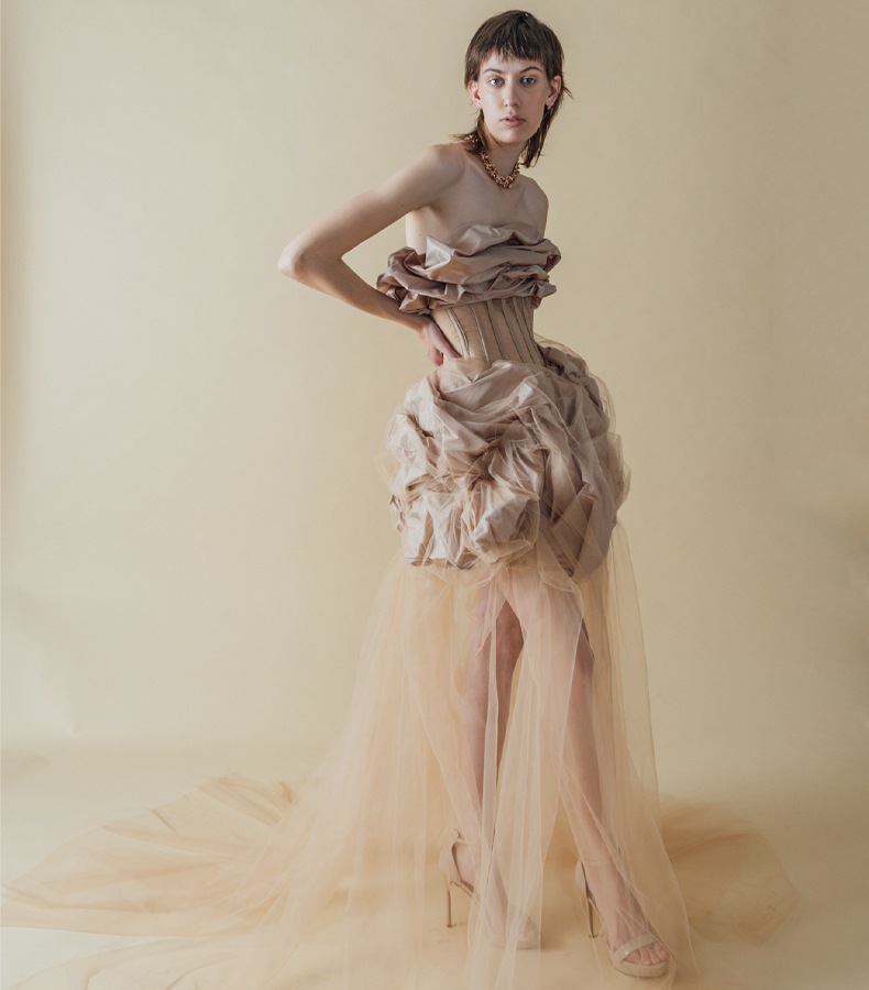 Photo of a model in a front three-quarter pose, wearing a blush beige silk taffeta gown that has taffeta draped on top, stopping just below the bust, and also draped at the skirt, starting just below the natural waist and stopping above the knee. The silk taffeta has a soft shine to it. Between the two draped sections is a very fitted, sheer beige organza paneled bodice, with beige boning channels to contrast the large, organic shapes on top and bottom. Draped with the silk taffeta on the skirt is a layer of beige netting, which is sewn down and tucked with the taffeta. The netting continues down past the bottom of the draped taffeta of the skirt and extends to the floor, creating a long, sheer skirt. The model wears nude shoes and a handmade, triple-strand choker made of beige and copper-toned organically shaped pearls. The model's right leg is slightly forward, crossed in front of her left leg, and her right elbow extends outward, with her right hand resting on her right hip. The background is a warmer-toned beige.