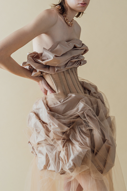 Photo of a model in a front three-quarter pose, wearing a blush beige silk taffeta gown that has taffeta draped on top, stopping just below the bust, and also draped at the skirt, starting just below the natural waist and stopping above the knee. The silk taffeta has a soft shine to it. Between the two draped sections is a very fitted, sheer beige organza paneled bodice, with beige boning channels to contrast the large, organic shapes on top and bottom. Draped with the silk taffeta on the skirt is a layer of beige netting, which is sewn down and tucked with the taffeta. The model wears a handmade, triple-strand choker made of beige and copper-toned organically shaped pearls. The model's right elbow is bent outward, with her right hand resting on her right hip. The background is a warmer-toned beige.