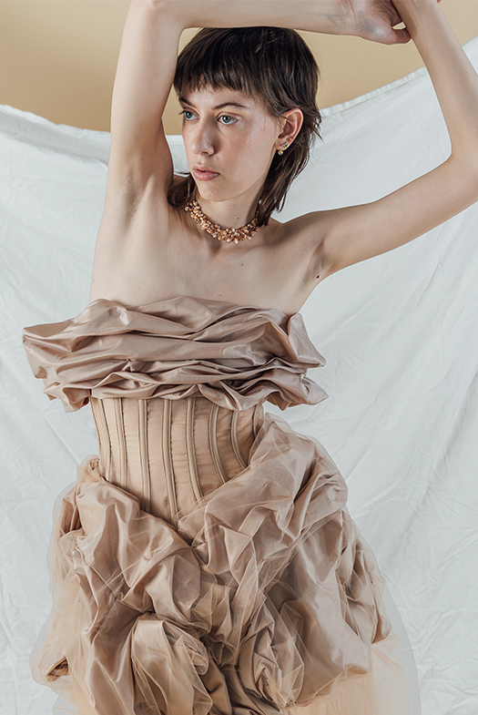 Photo of a model in a front pose, arms over her head and with her right hand holding her left wrist, wearing a blush beige silk taffeta gown that has taffeta draped on top, stopping just below the bust, and also draped at the skirt, starting just below the natural waist. The silk taffeta has a soft shine to it. Between the two draped sections is a very fitted, sheer beige organza paneled bodice, with beige boning channels to contrast the large, organic shapes on top and bottom. Draped with the silk taffeta on the skirt is a layer of beige netting, which is sewn down and tucked with the taffeta as an overlay. The model wears a handmade, triple-strand choker made of beige and copper-toned organically shaped pearls. The background is a warmer-toned beige with a white sheet draped partially across it.