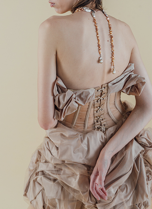 Photo of a model in a close-up back pose, with her left arm next to her body, her left hand in front of her, her right arm bent around her back, and her right hand resting palm-up on the back of the skirt. She's wearing a blush beige silk taffeta gown that has taffeta draped on top, stopping just below the bust and getting thinner towards the center back, and also draped at the skirt, starting just below the natural waist. The silk taffeta has a soft shine to it. Between the two draped sections is a very fitted, sheer beige organza paneled bodice, with beige boning channels to contrast the large, organic shapes on top and bottom. At the center back, there are antique copper grommets, and the corset bodice laces up with a light beige suede string. Draped with the silk taffeta on the skirt is a layer of beige netting, which is sewn down and tucked with the taffeta as an overlay. The model wears a handmade, triple-strand choker made of beige and copper-toned organically shaped pearls. Hanging from the back of the necklace are two separate strands of small copper and beige-toned pearls, each with a large baroque pearl at the end, resting right above the back of the dress. The background is a warmer-toned beige.