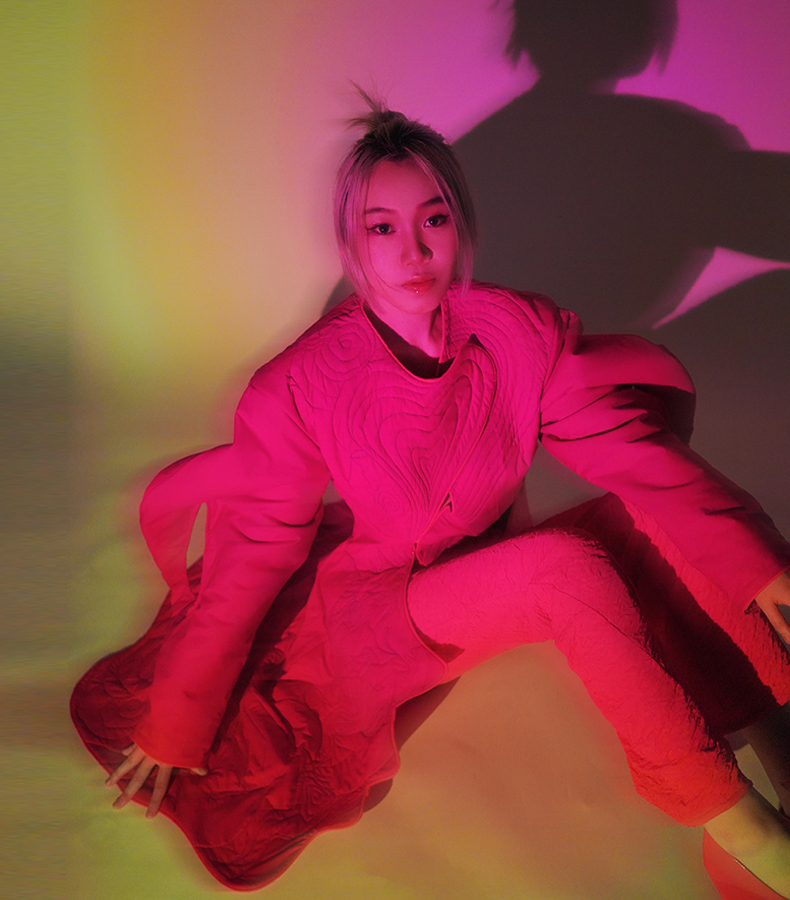 The model sits on the ground, wearing a Barbie pink quilted coat and hot pink pants. There is a heart-shaped closure at the front. The heart shape is quilted with lines.