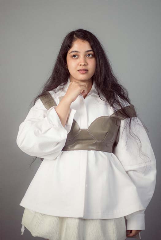Portrait view of a girl wearing an oversized shirt under a bralette. She is posing with her right hand on her chin with an unbuttoned cuff. The shirt is flared out at the hem, which displays the short white bubble skirt. 
