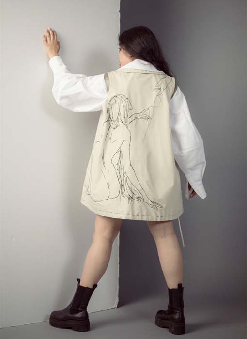 Girl posing with her left hand on the wall showing the back of the embroidered vest. The color of the vest is off-white with black stitching. The art demonstrates a girl sitting and reaching her hand out to another. The embroidery includes loose threads hanging off the vest at the girl's hair and hands. 