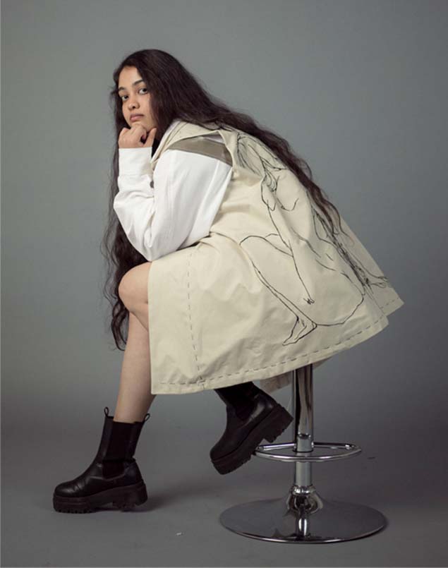 The girl posed sitting with a side view with one hand resting on her knee and her chin placed on her hand. The image further shows the embroidery around the vest, with thread tracing detailing the hem. 