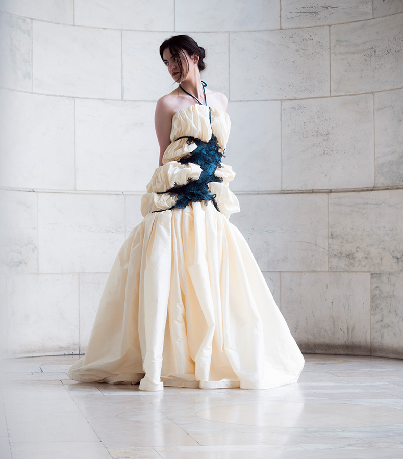 A woman in an alcove at the New York Public Library. She is wearing an ivory-covered dress that forms large poufs. The dress is constrained by a teal piece with layers of overlapping lace. Cords wrap around the body, containing the poufs.