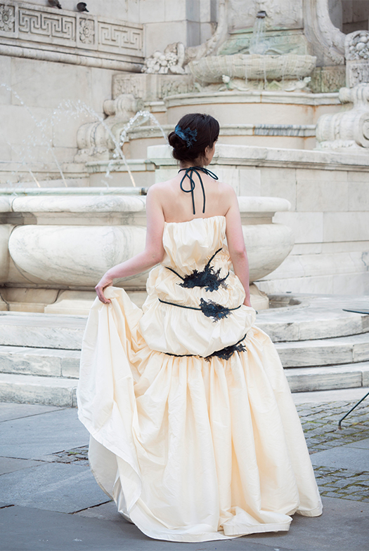 A view of the back of the gown. The skirt is intensely gathered and hangs from mid-hip. The hem drapes over the ground. The back of the dress has several pieces of lace that join the cords together.