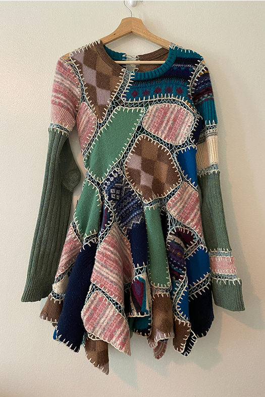 Full view of dress on a hanger, white background. Dress is made of patchwork sweaters in many patterns and colors with the patterns facing many directions. It has a full skirt, crew neck, and long sleeves made from long green socks.